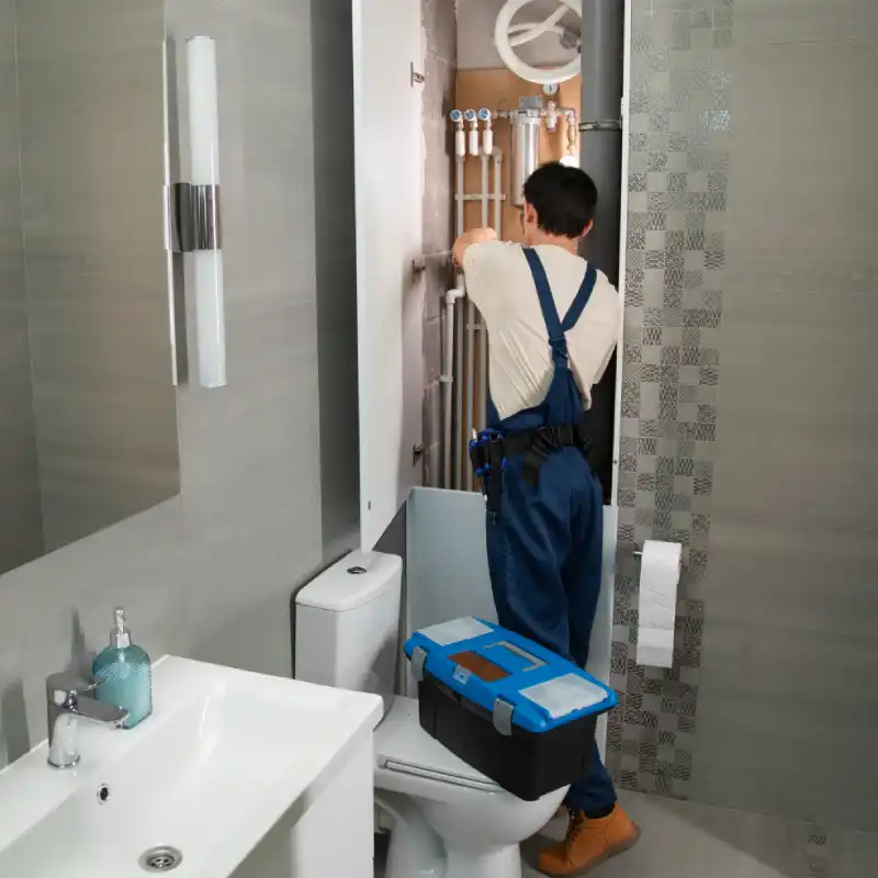 A plumber in a blue jumpsuit works on pipes behind a cabinet in a modern bathroom, with tools on the toilet nearby